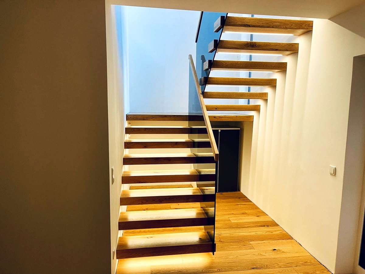 Kragarmtreppe mit Stufen aus Eiche, unter jeder Stufe eine Lichtleiste, daneben ein Glasgeländer mit Handlauf aus Holz