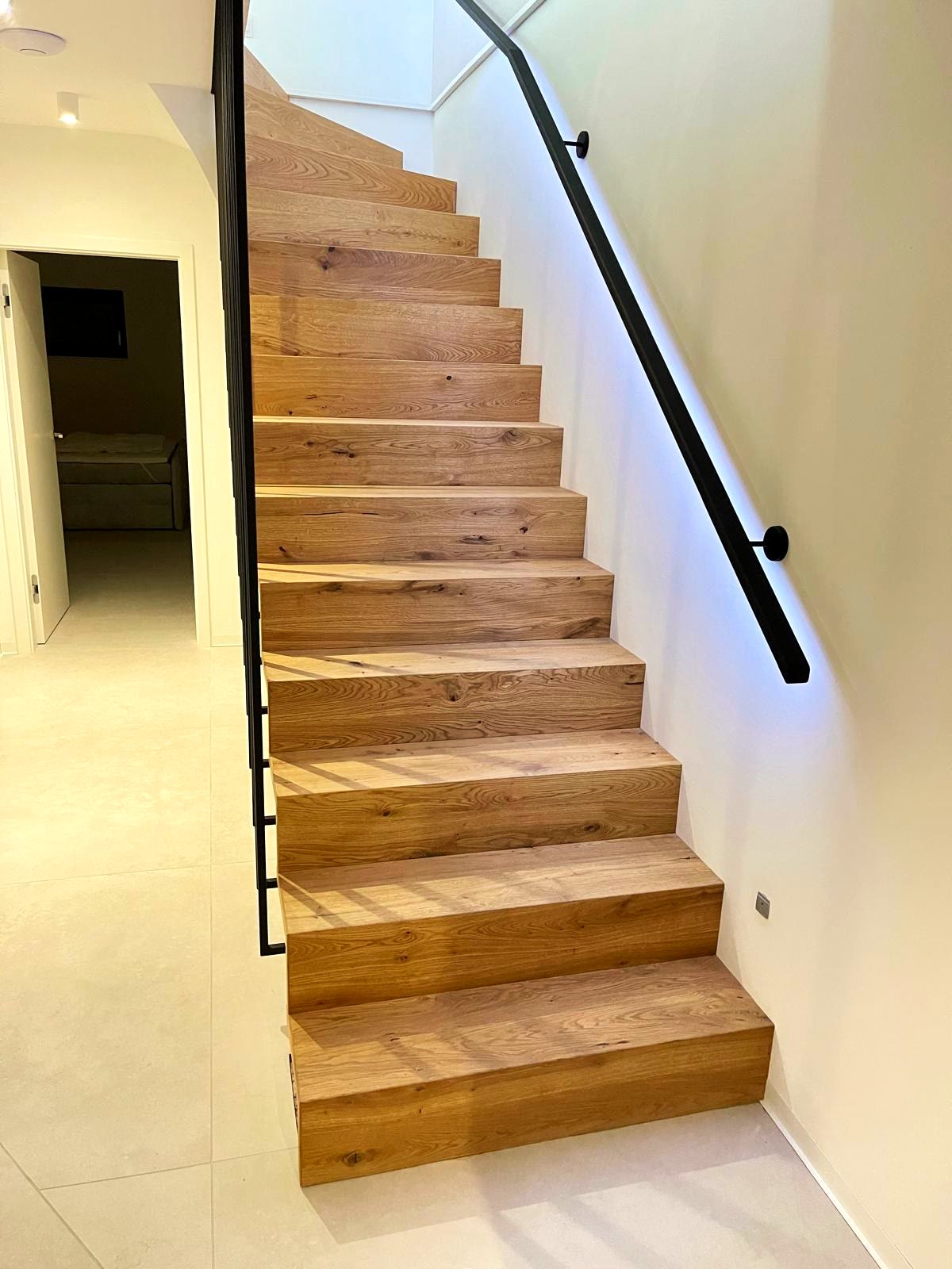 Betontreppe mit Eichenstufen belegt, schwarzer Handlauf an der Wand, indirekte Beleuchtung dahinter