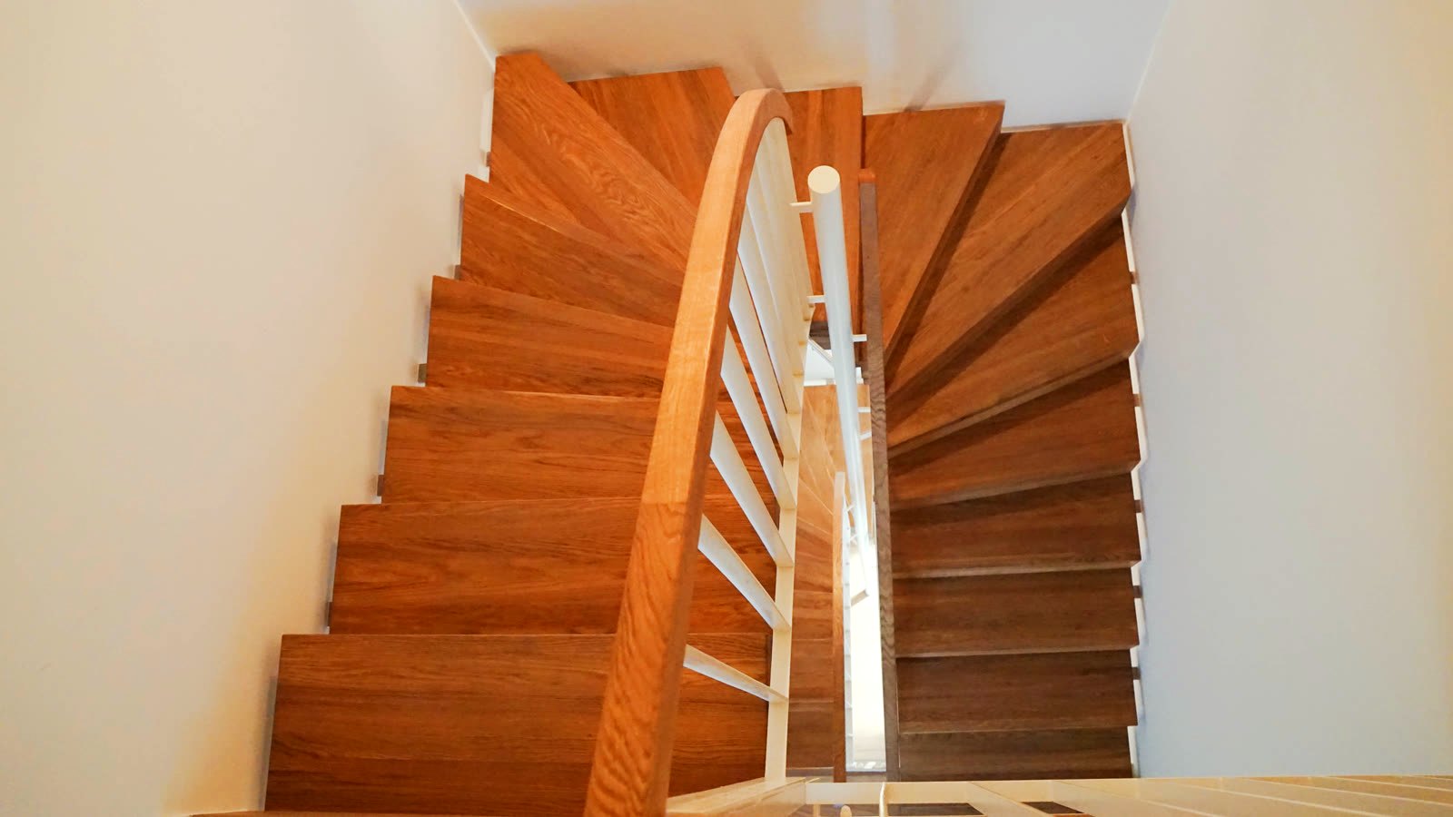 Blick von oben auf eine gewendelte Massivholztreppe mit weißem Stabgeländer und geschwungenem Holzhandlauf